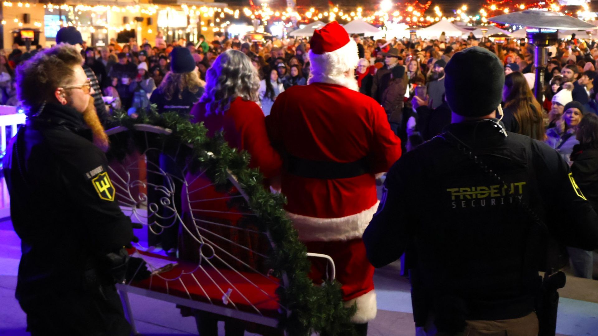 Santa Claus and a person in a red outfit stand before a large crowd at an outdoor evening holiday event with security.
