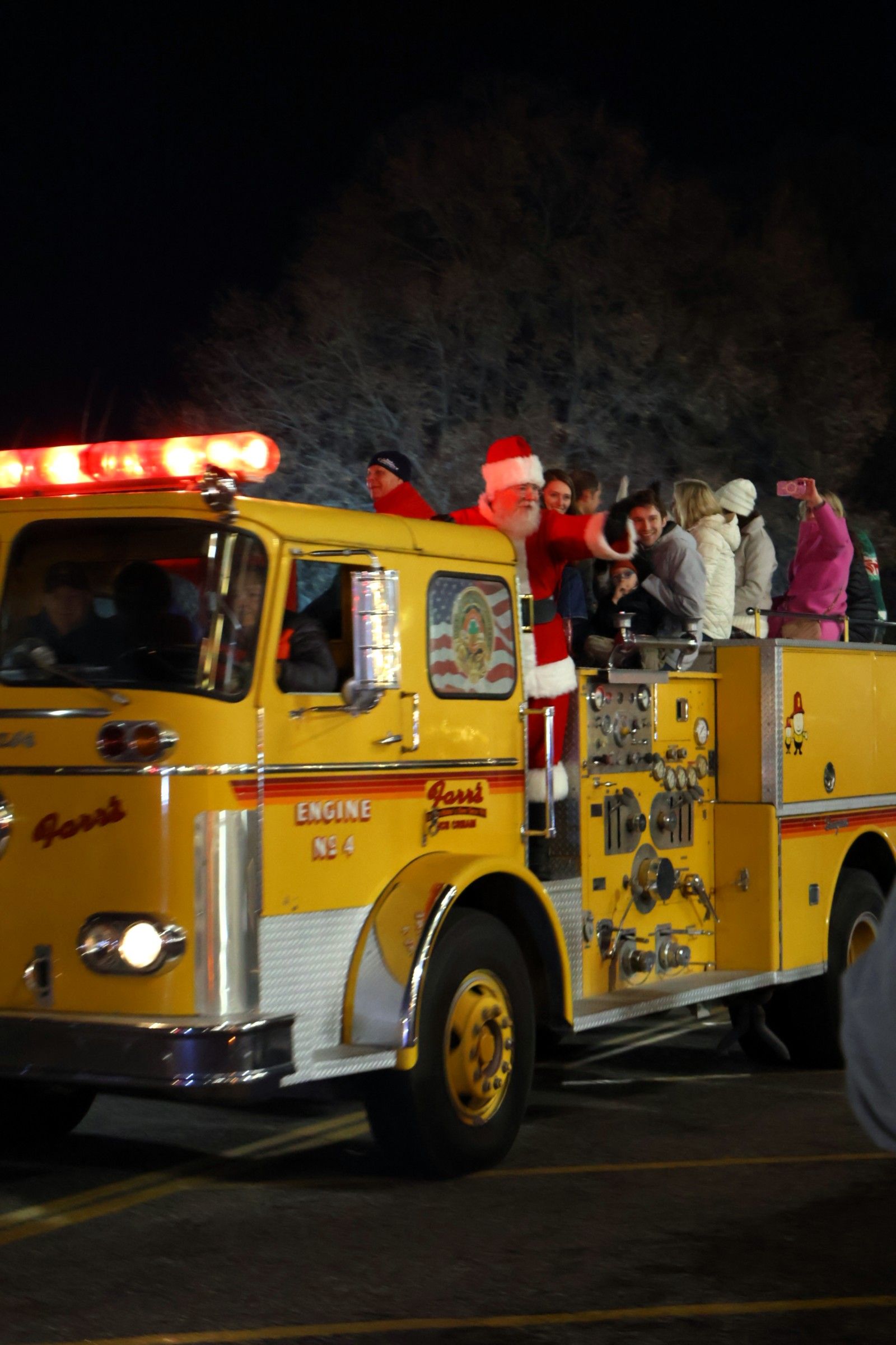 Santa Claus waves from atop a bright yellow fire engine during a nighttime community parade.