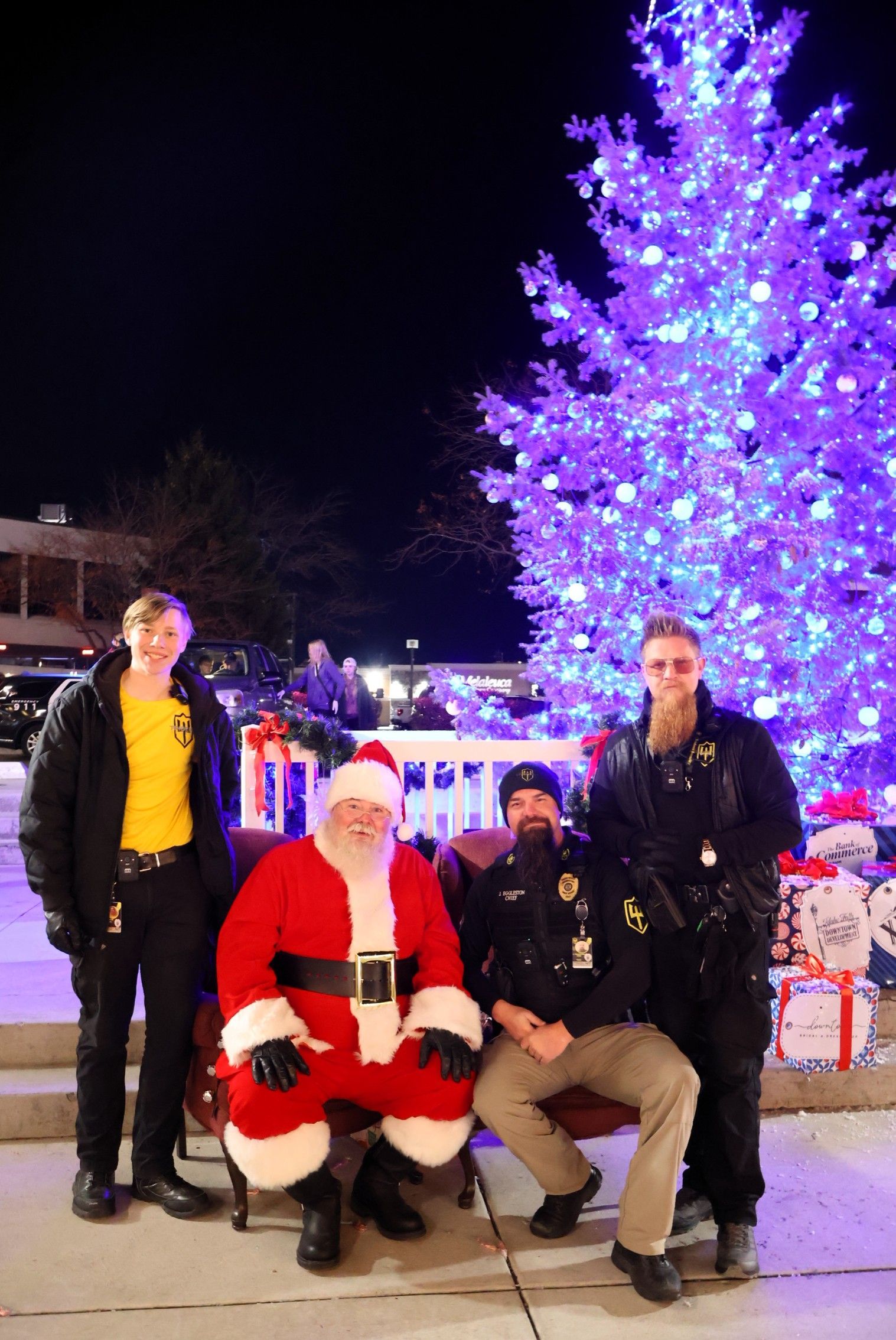 Santa Claus sits on a bench with two uniformed officers in front of a large, brightly lit purple Christmas tree at night.