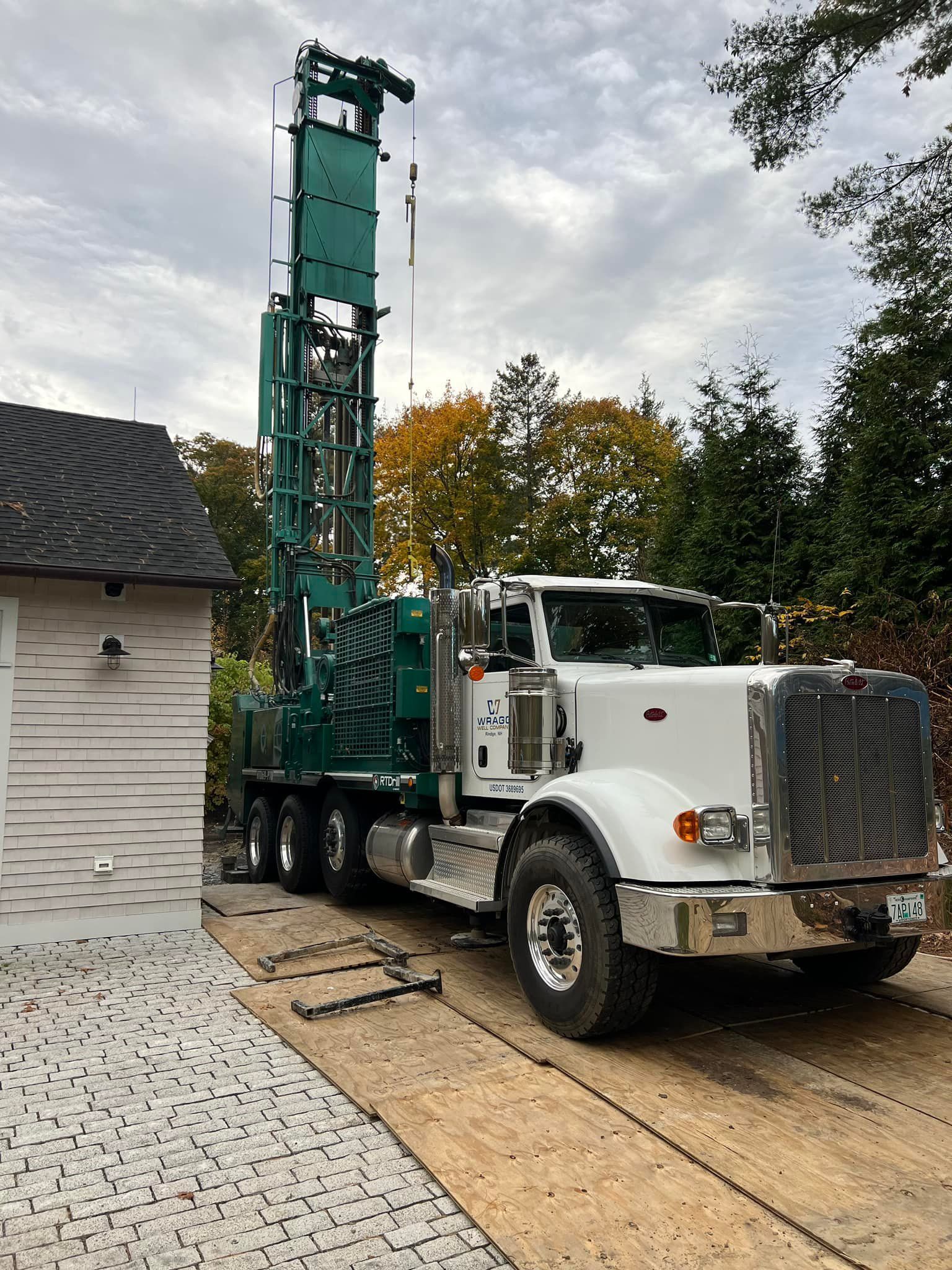 A white semi truck with a green drilling rig on the back is parked in front of a garage.
