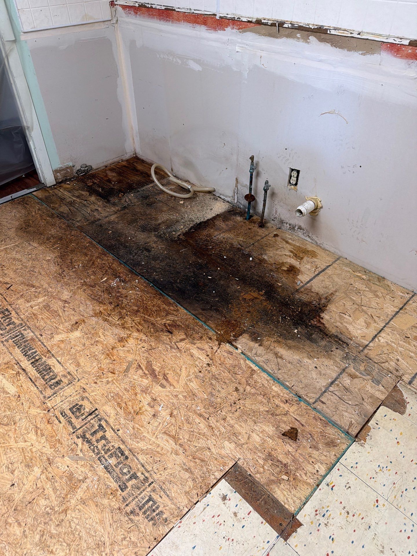 Water-damaged floor with dark stain and exposed subfloor beside a wall outlet opening