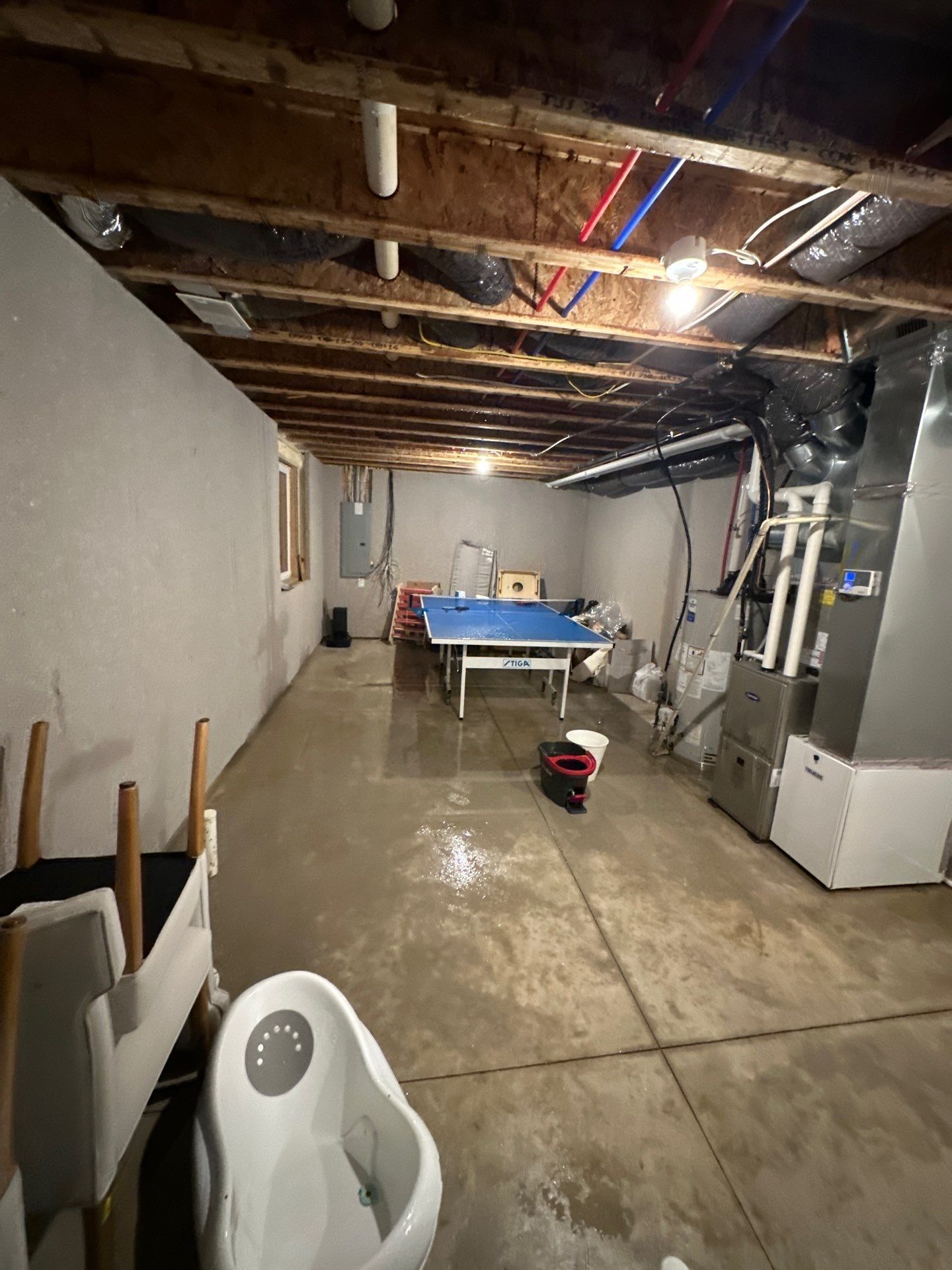 Basement with exposed ceiling beams, concrete floor, workbench, and furnace in a utility/storage area.