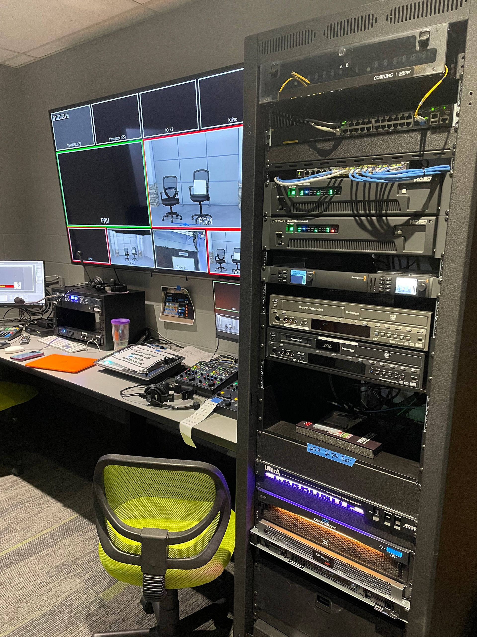A room with a desk , chair , monitors and a server rack.