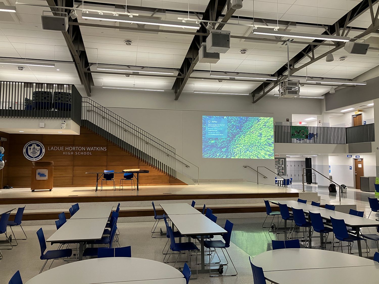 A large room with tables and chairs and a large screen on the wall.