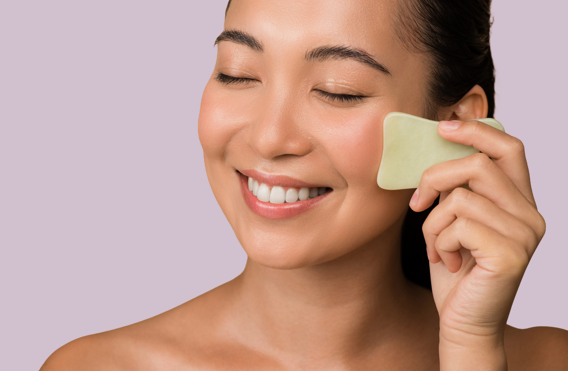 Woman with closed eyes smiling, using a jade gua sha tool on her cheek, lavender background.