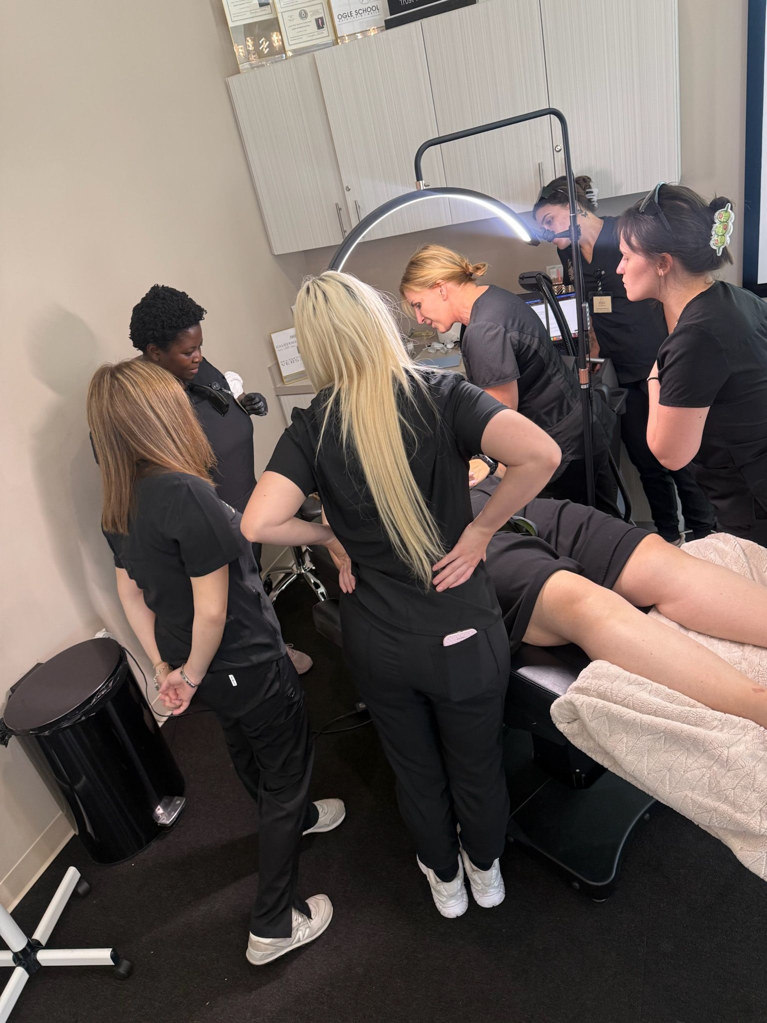 Aesthetics students observe a hands-on treatment demonstration around a client at a training academy in Coppell, TX