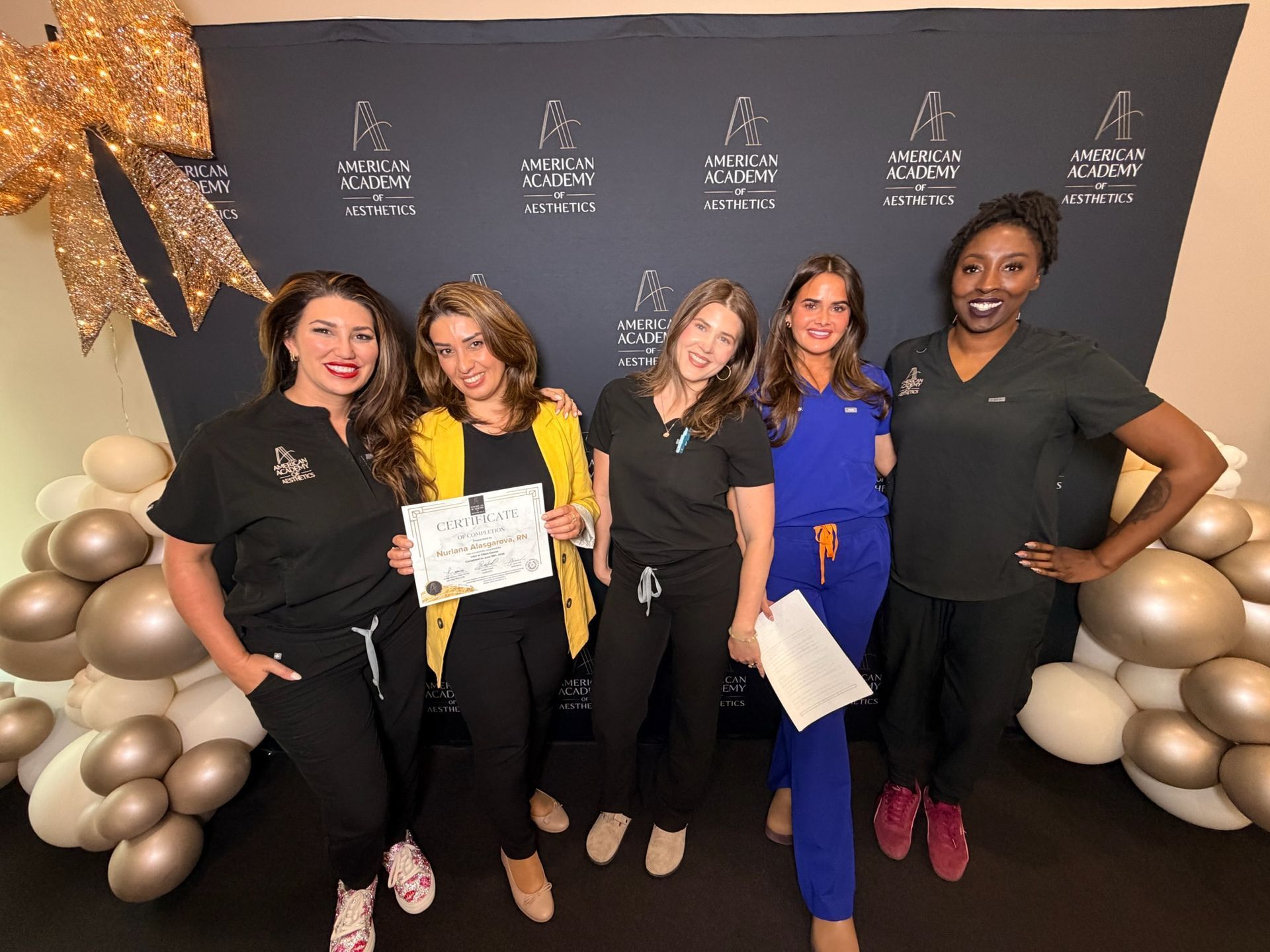 Five women posing at a celebration backdrop with gold and silver balloons and one holding a certificate