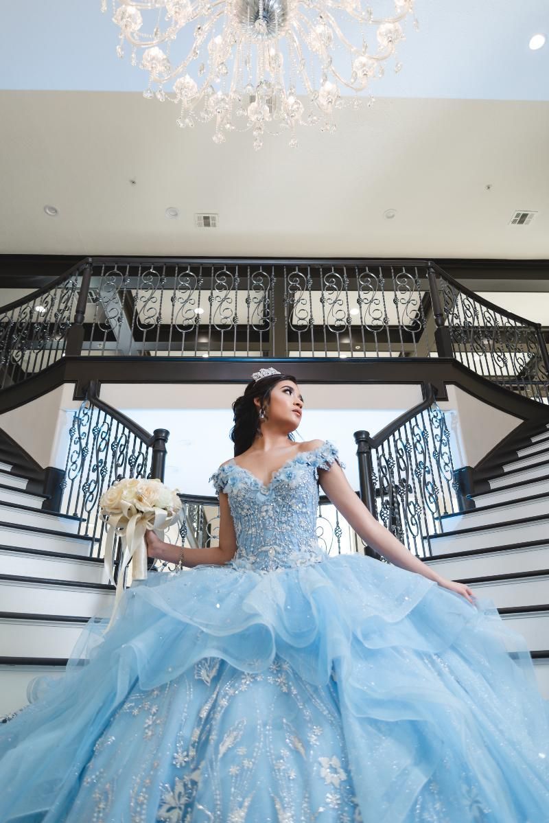 A woman in a blue ball gown is standing on a set of stairs holding a bouquet of flowers.