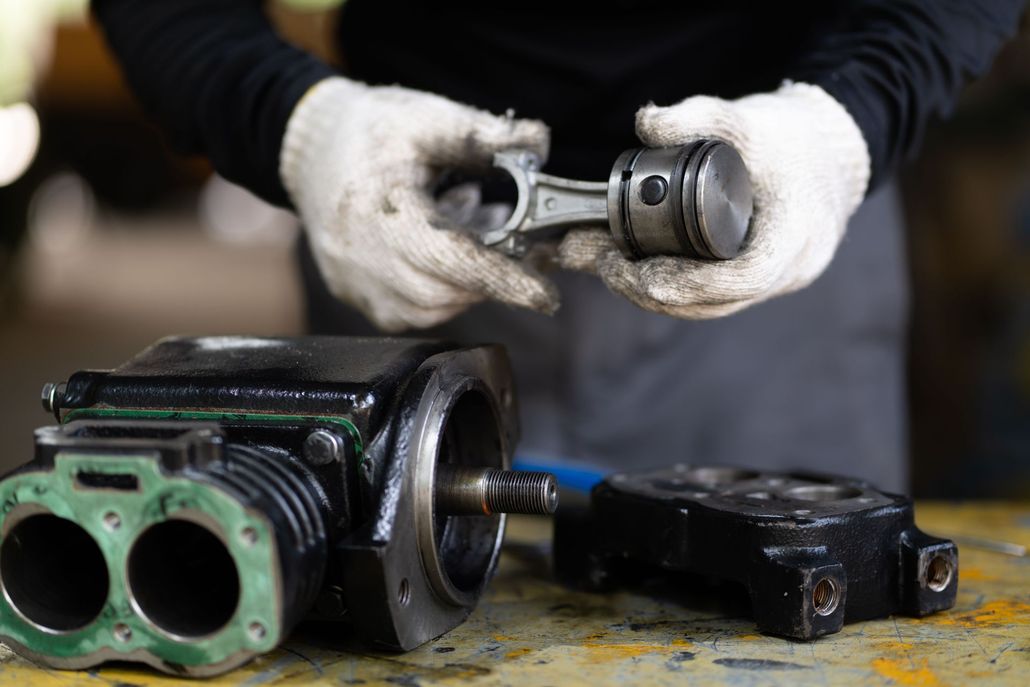 Gloved hands holding a metal engine part above two disassembled black-green mechanical components on a workbench
