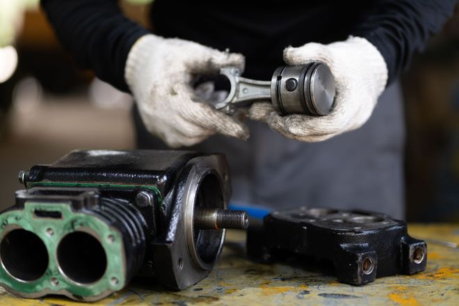 Gloved hands holding a metal engine part above two disassembled black-green mechanical components on a workbench