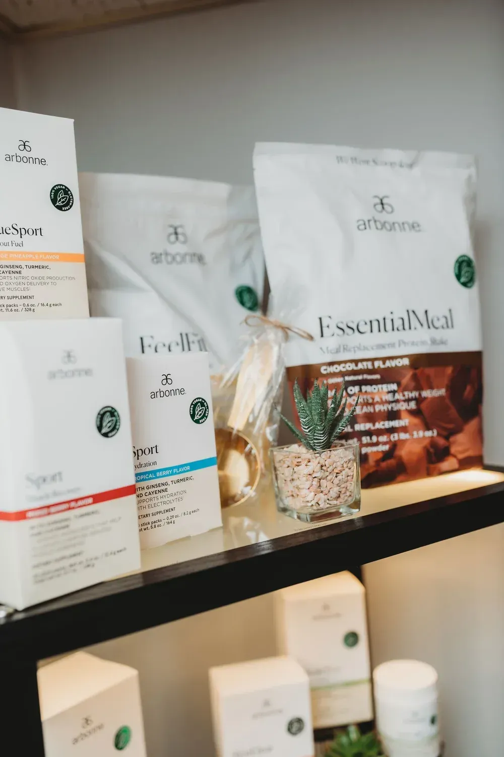 Arbonne nutritional products, including pouches and boxes, are displayed on shelves with a small succulent in a pot.