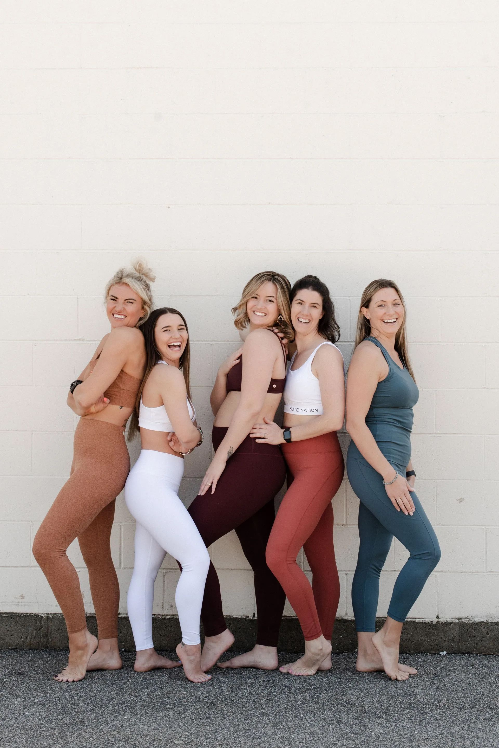 Five women posing together against a white wall, wearing colorful matching outfits and smiling.