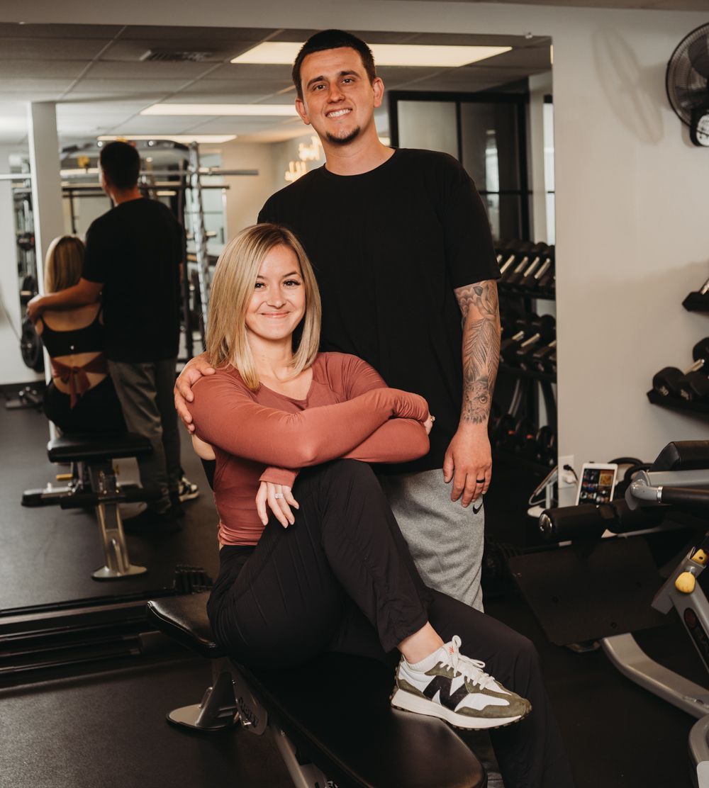 Two people posing in a gym, one seated and one standing, with workout equipment in the background