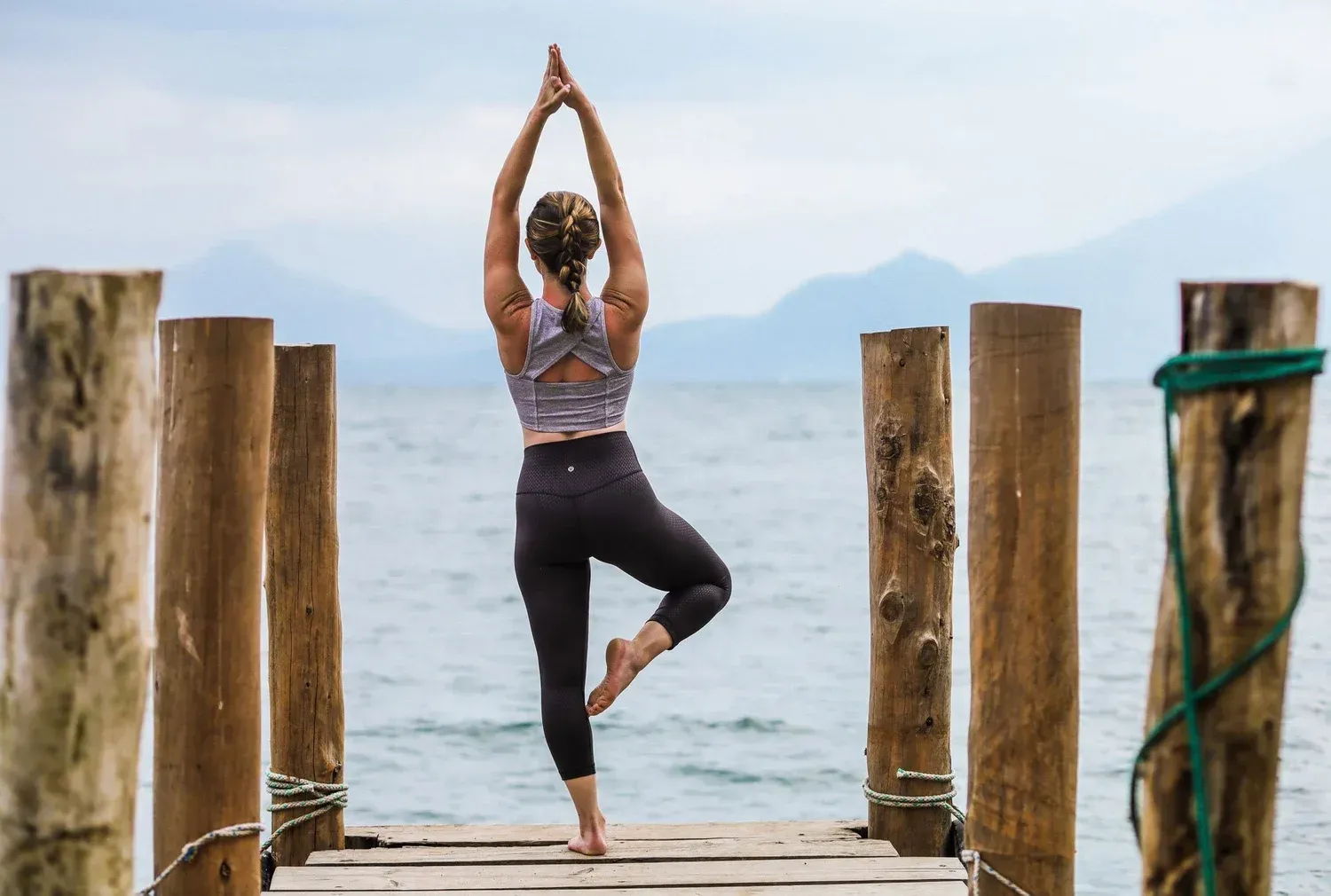 Woman in yoga tree pose on wooden dock, arms raised, facing a lake and mountains.