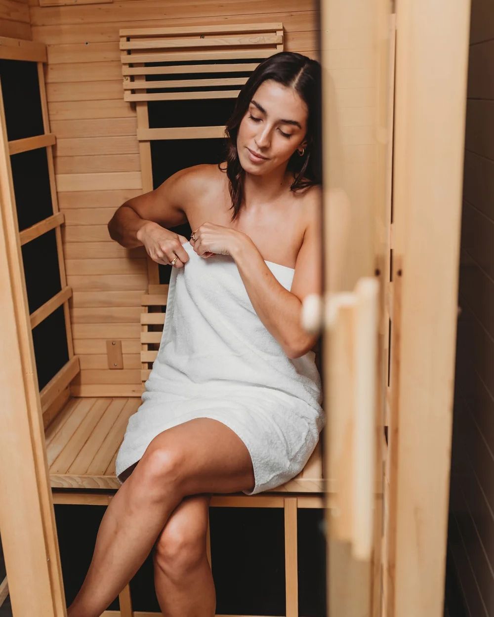 Woman wrapped in a towel, seated in a wooden sauna, smiling.