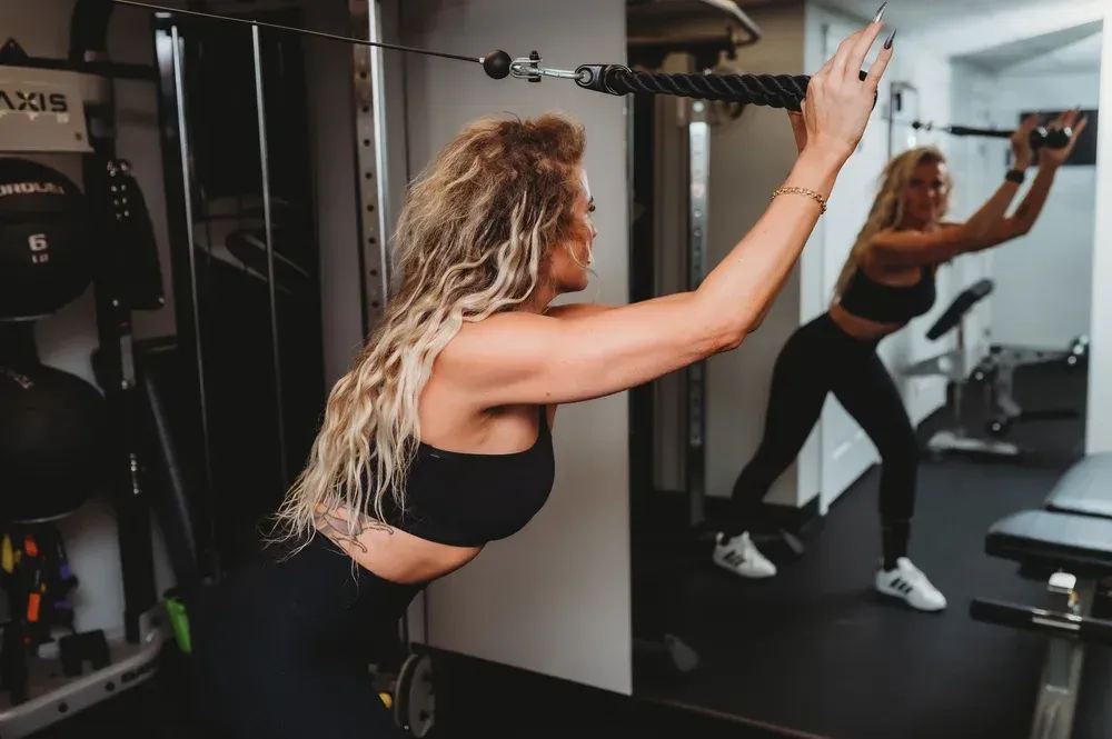 Woman working out at a gym, pulling cable towards herself. Black workout clothes, mirror reflection.