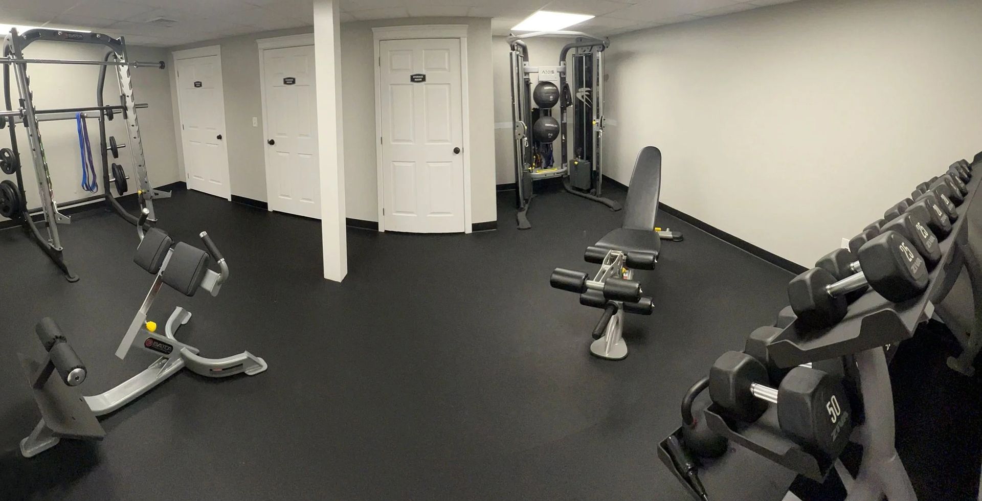Gym with weight rack, machines, dumbbells, and benches on a black floor, with white walls and doors.