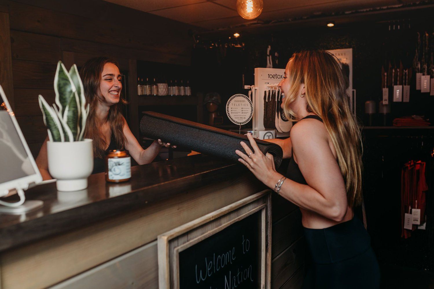 Woman at a counter receiving a rolled-up yoga mat from a staff member. Dark interior, neutral colors.