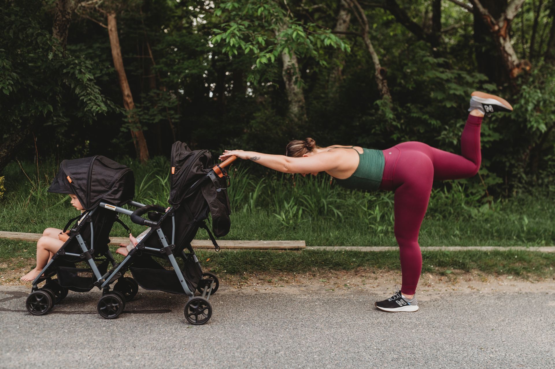 Woman doing yoga pose, leg extended, while holding stroller handle. Forest setting.