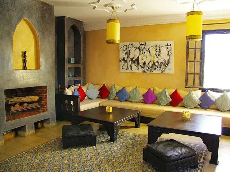 Cozy living room with fireplace, yellow walls, L-shaped sofa, colorful pillows, dark wooden tables, and hanging yellow lamps.
