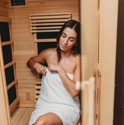 Woman wrapped in a white towel sitting inside a wooden sauna, adjusting her towel.