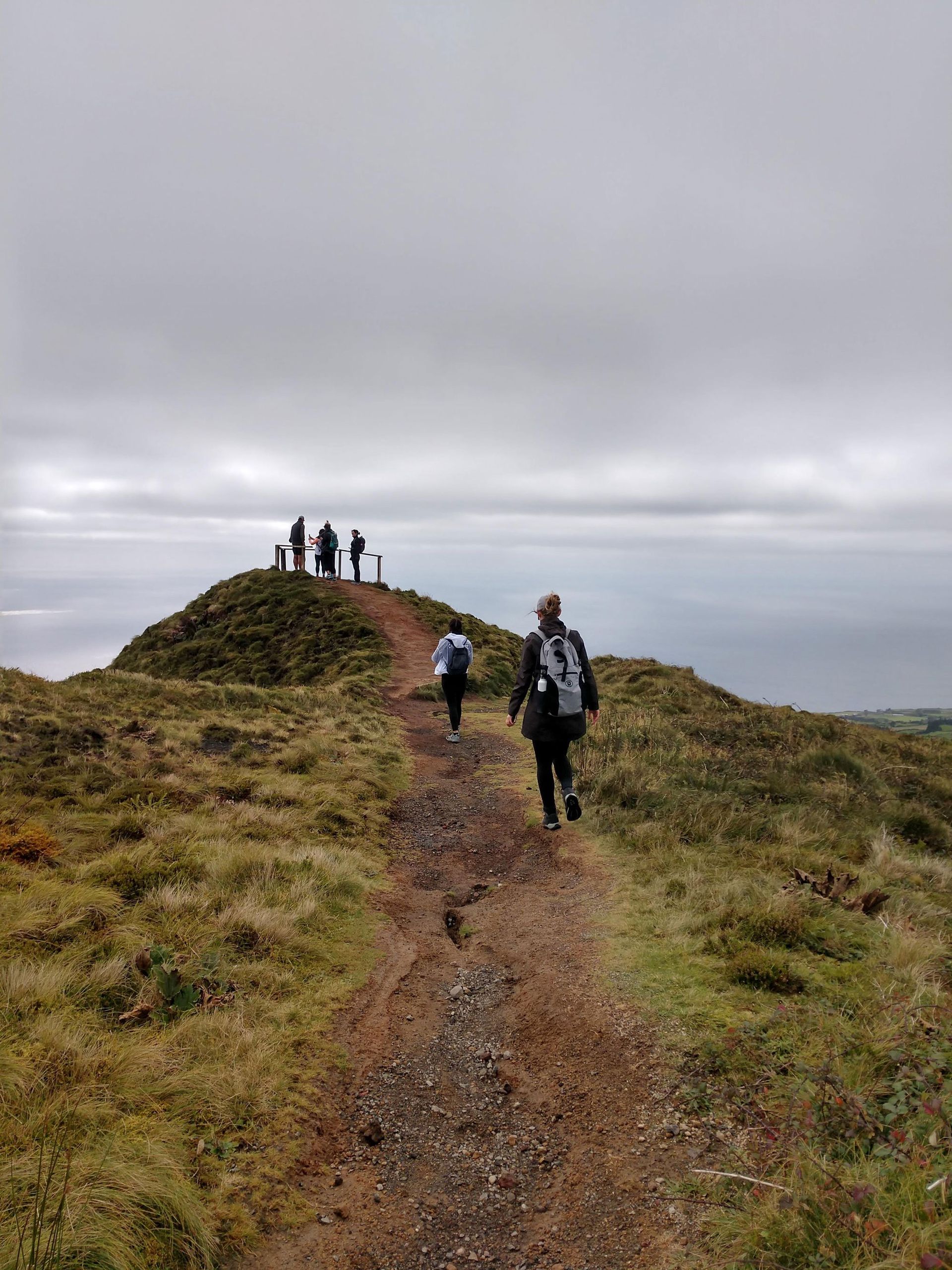 People hiking on a grassy trail toward a viewpoint with figures, under a cloudy sky.
