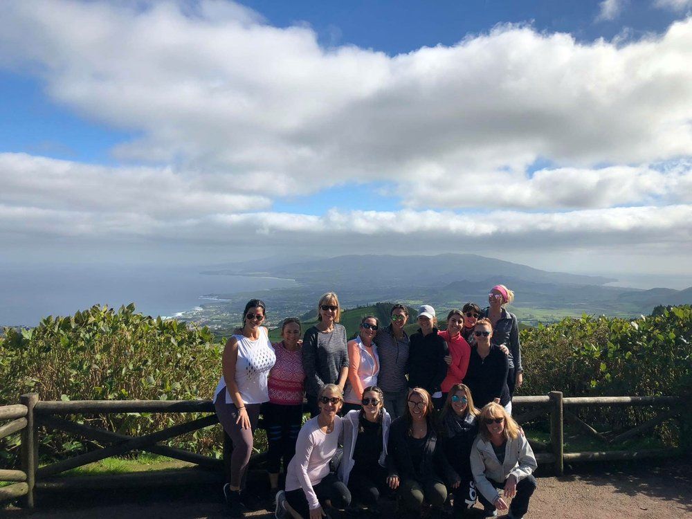 Group of people posing outdoors with a view of a coastline and mountains under a cloudy sky.
