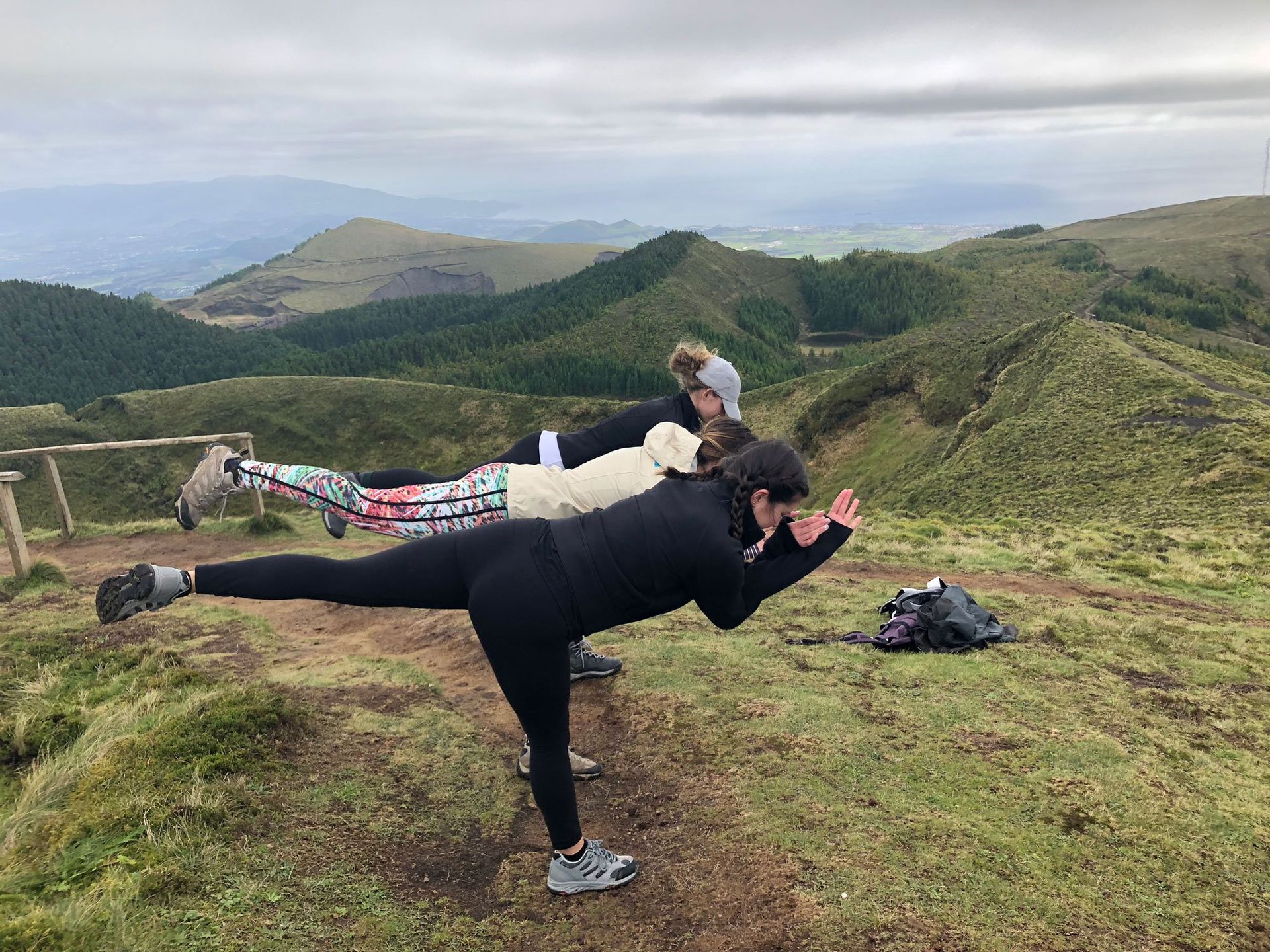 Three people doing a yoga pose on a mountaintop. Green hills and cloudy sky in background.