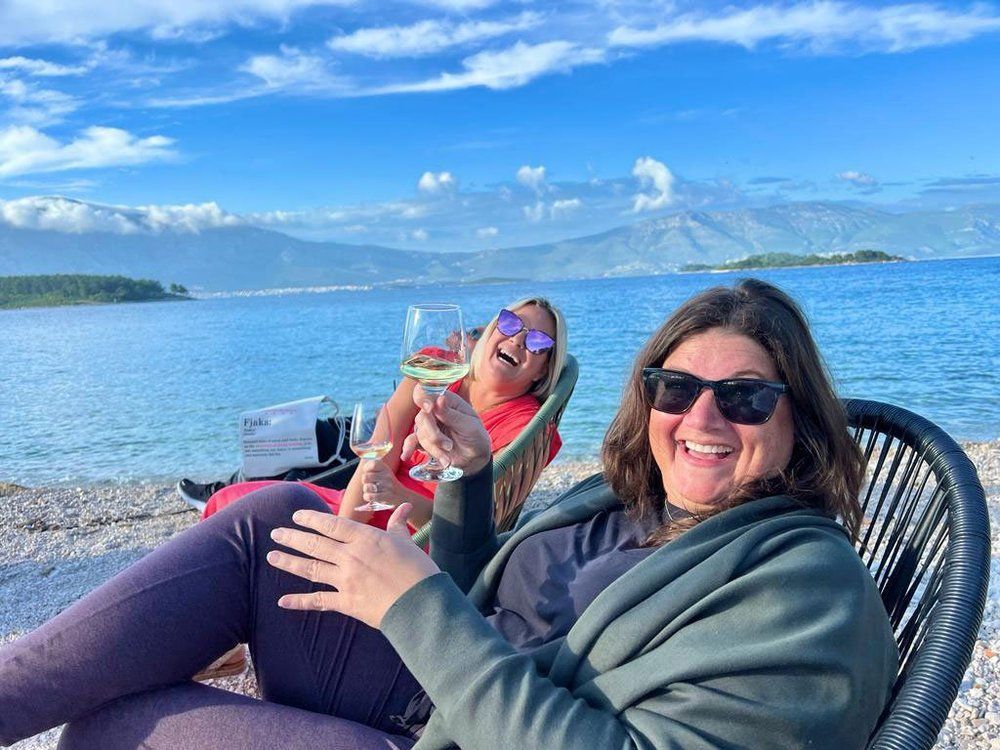 Two women laugh while drinking wine on a beach, mountains and sea in background.