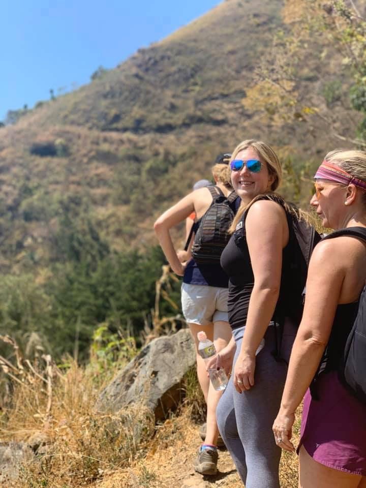 Three women hiking on a hillside trail, sunny day. One woman smiles, wears sunglasses.
