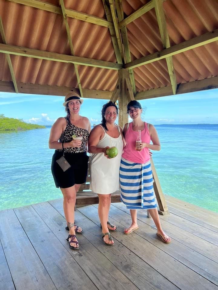 Three women stand on a wooden deck by the ocean, holding drinks. They smile in front of a blue sky and water.
