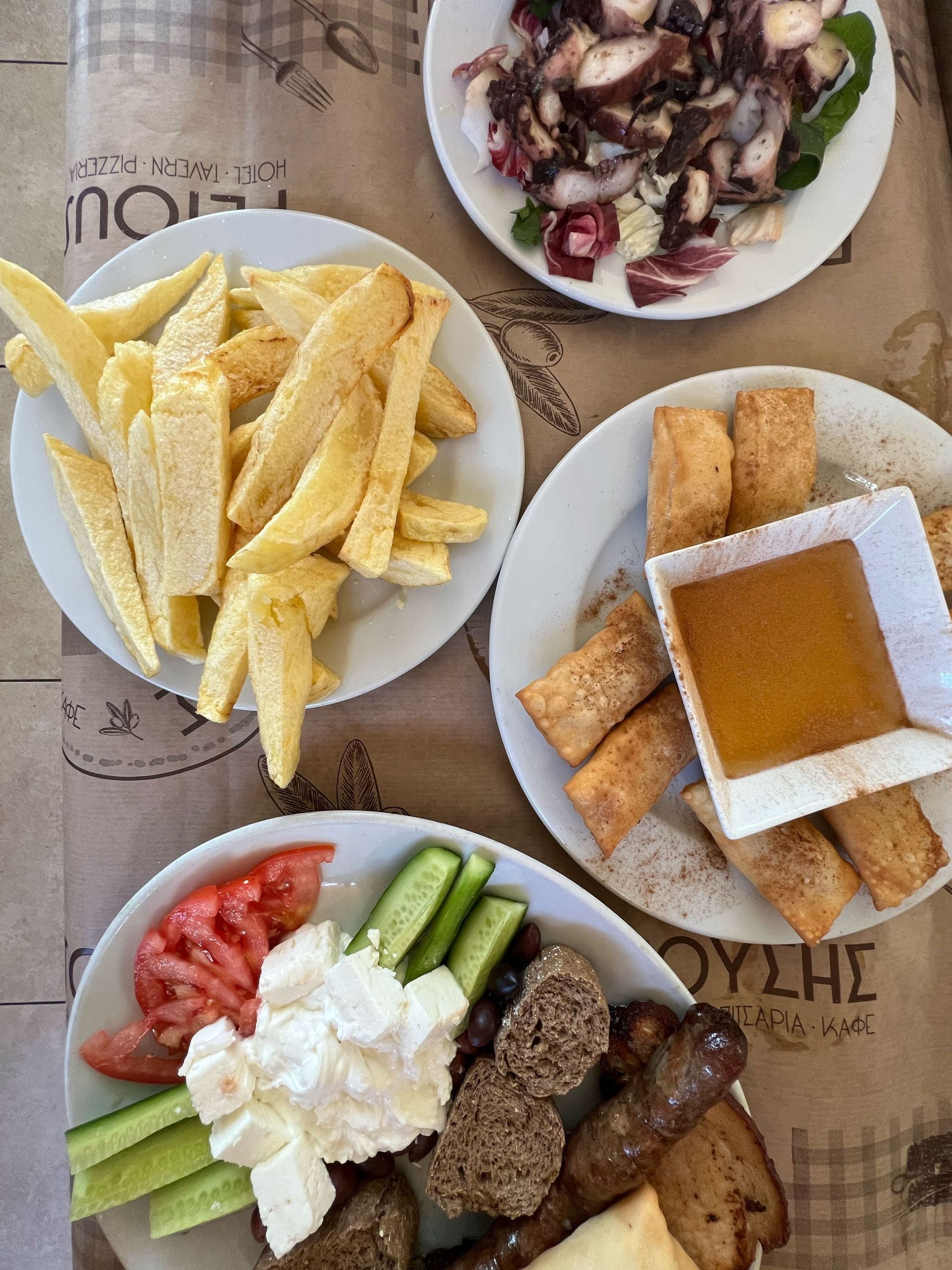 Plates of food: fries, octopus salad, cheese pastries with dipping sauce, and a plate with tomatoes, cucumbers, cheese, and sausage.