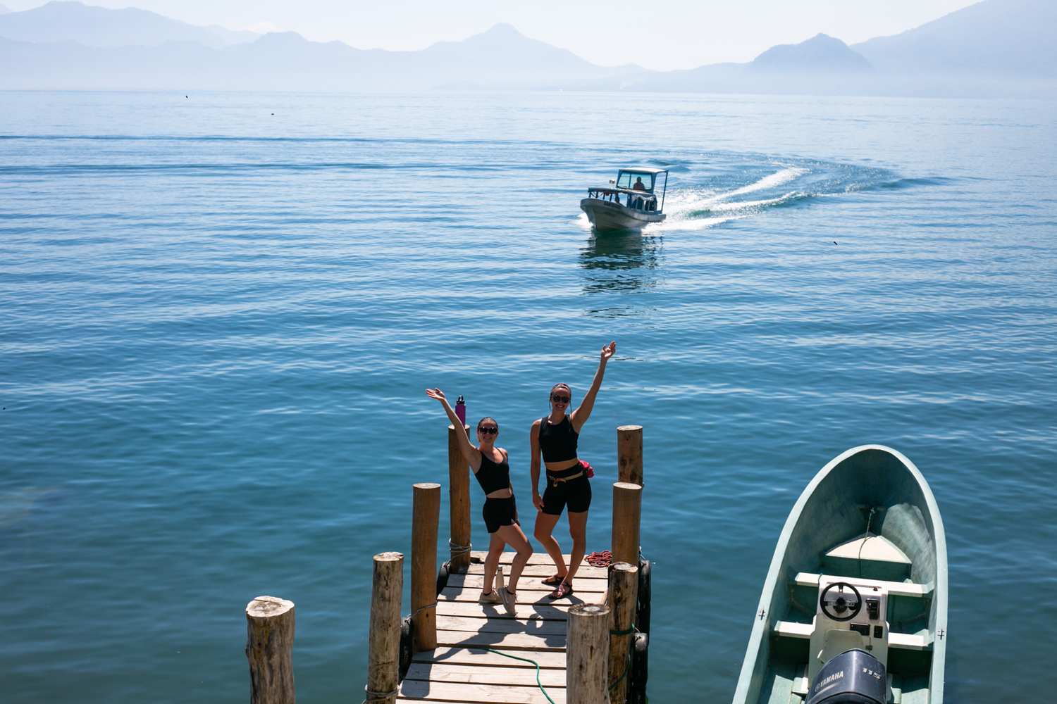 Two people wave from a wooden dock on a blue lake; a boat moves in the distance, a small boat is docked.