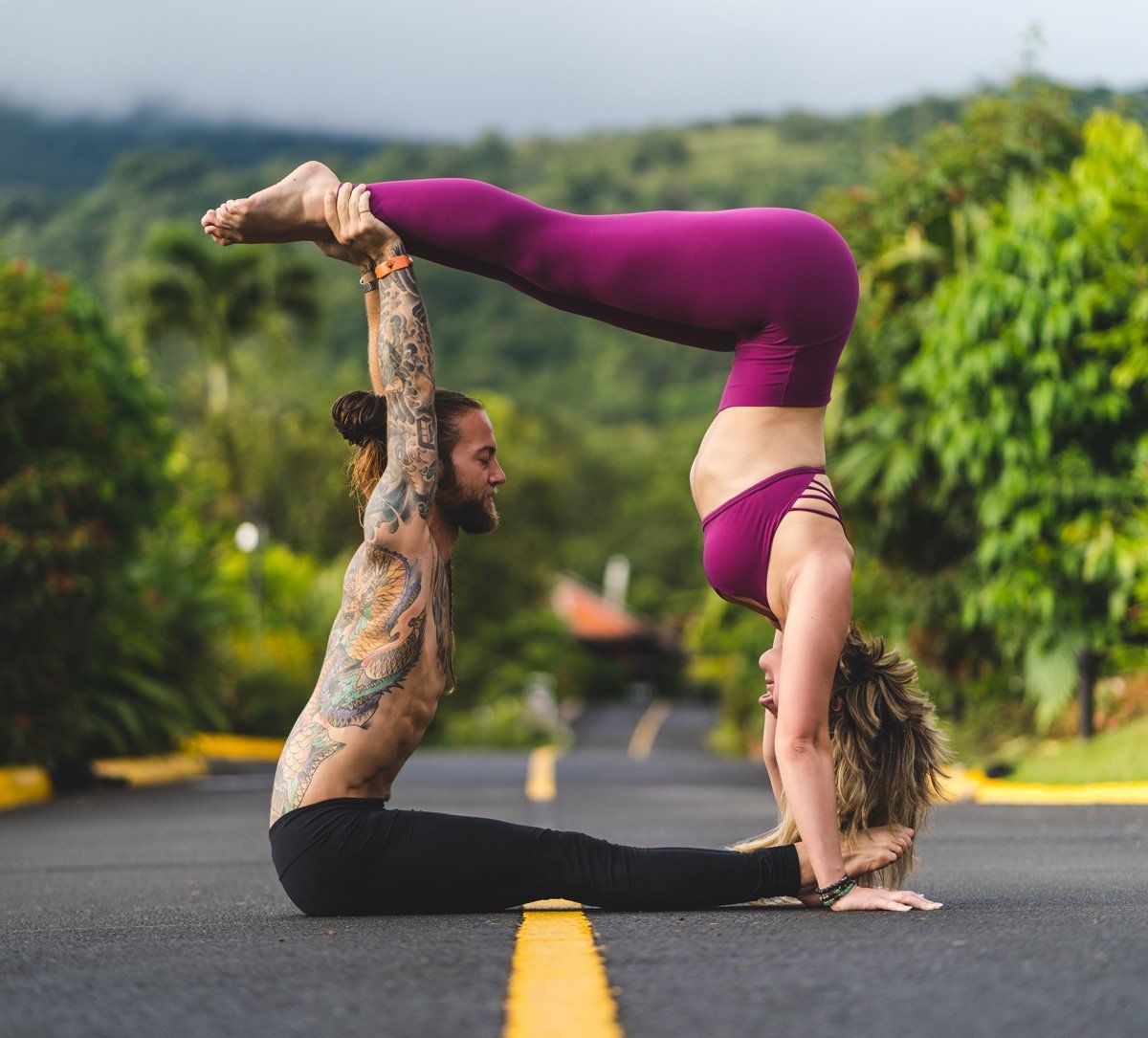 Two people perform acrobatic yoga on a road; one balances the other's pose with their feet.