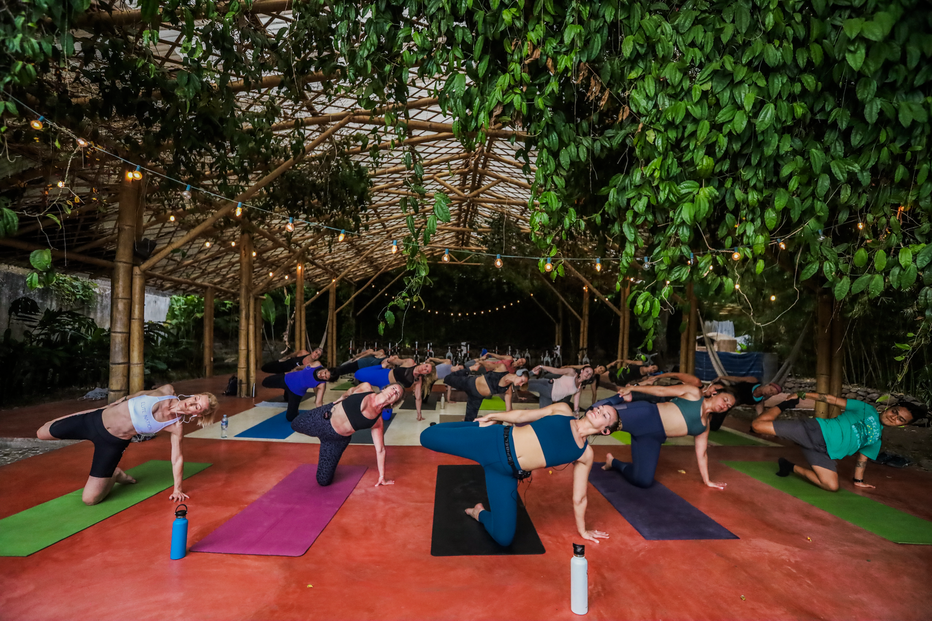 People in yoga poses under a bamboo and foliage canopy, outdoors on mats.
