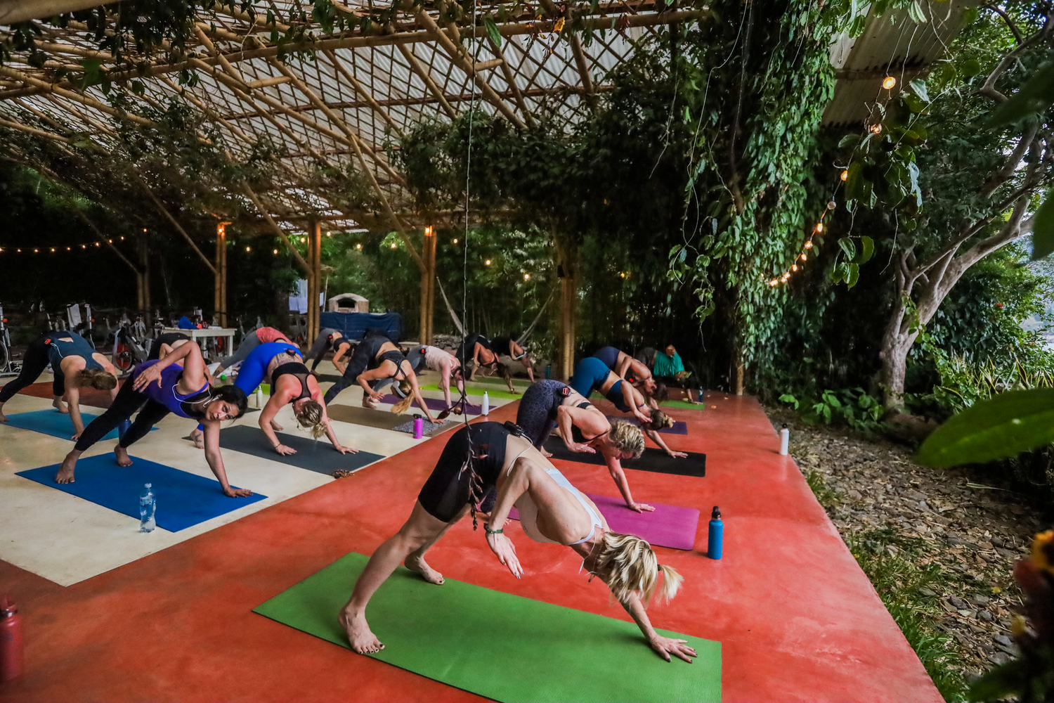 People doing yoga outdoors on a red-painted platform, under a latticed roof.