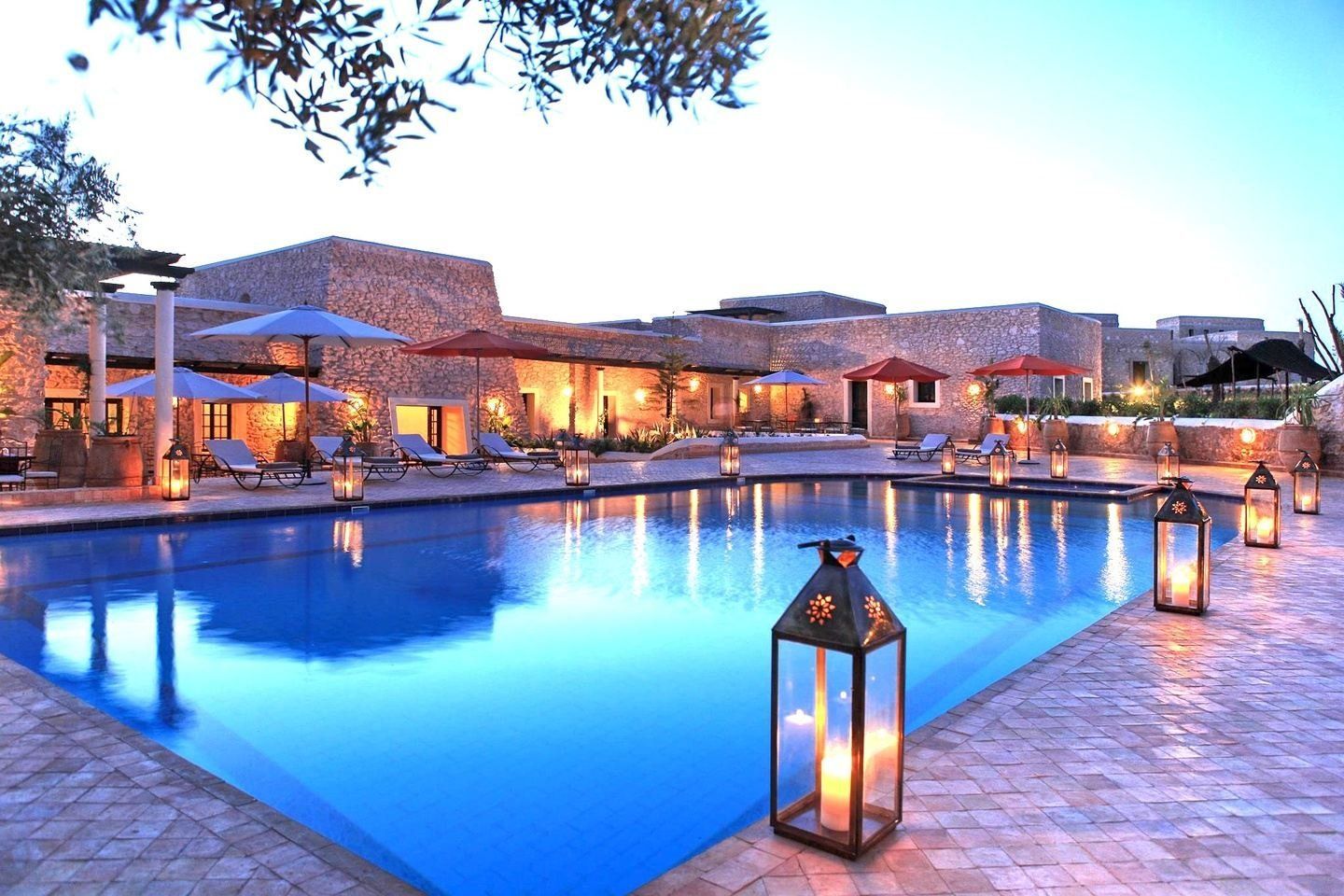 Pool at a resort with lanterns, lounge chairs, and buildings with stone walls.