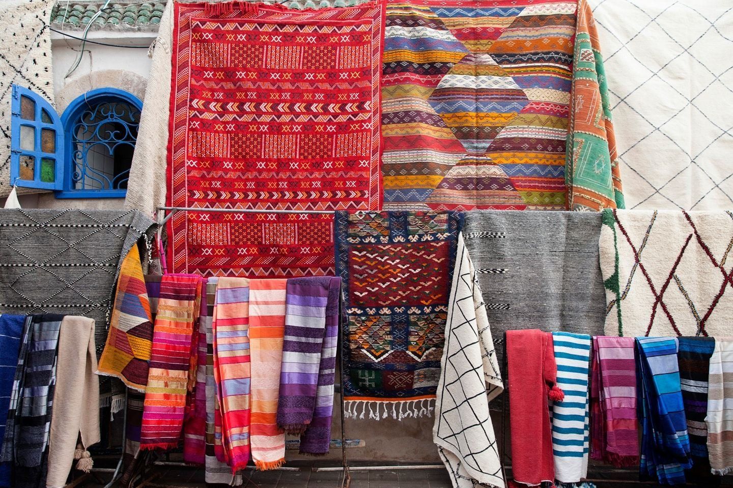 Colorful textiles hanging outdoors, including rugs and scarves, with a blue arched window in the background.