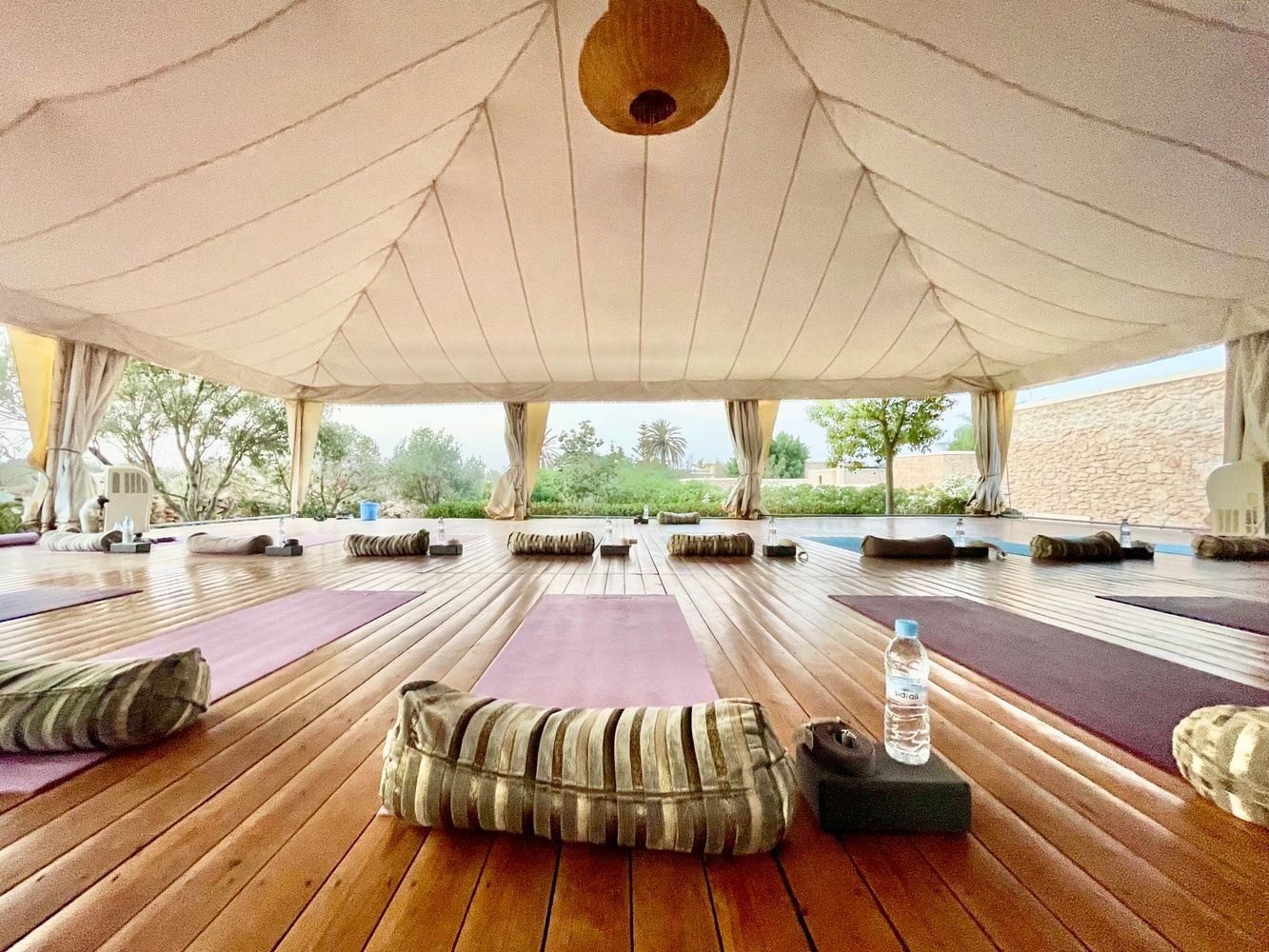 Yoga studio with purple mats, cushions, and wooden floor under a canvas roof, open to a garden view.