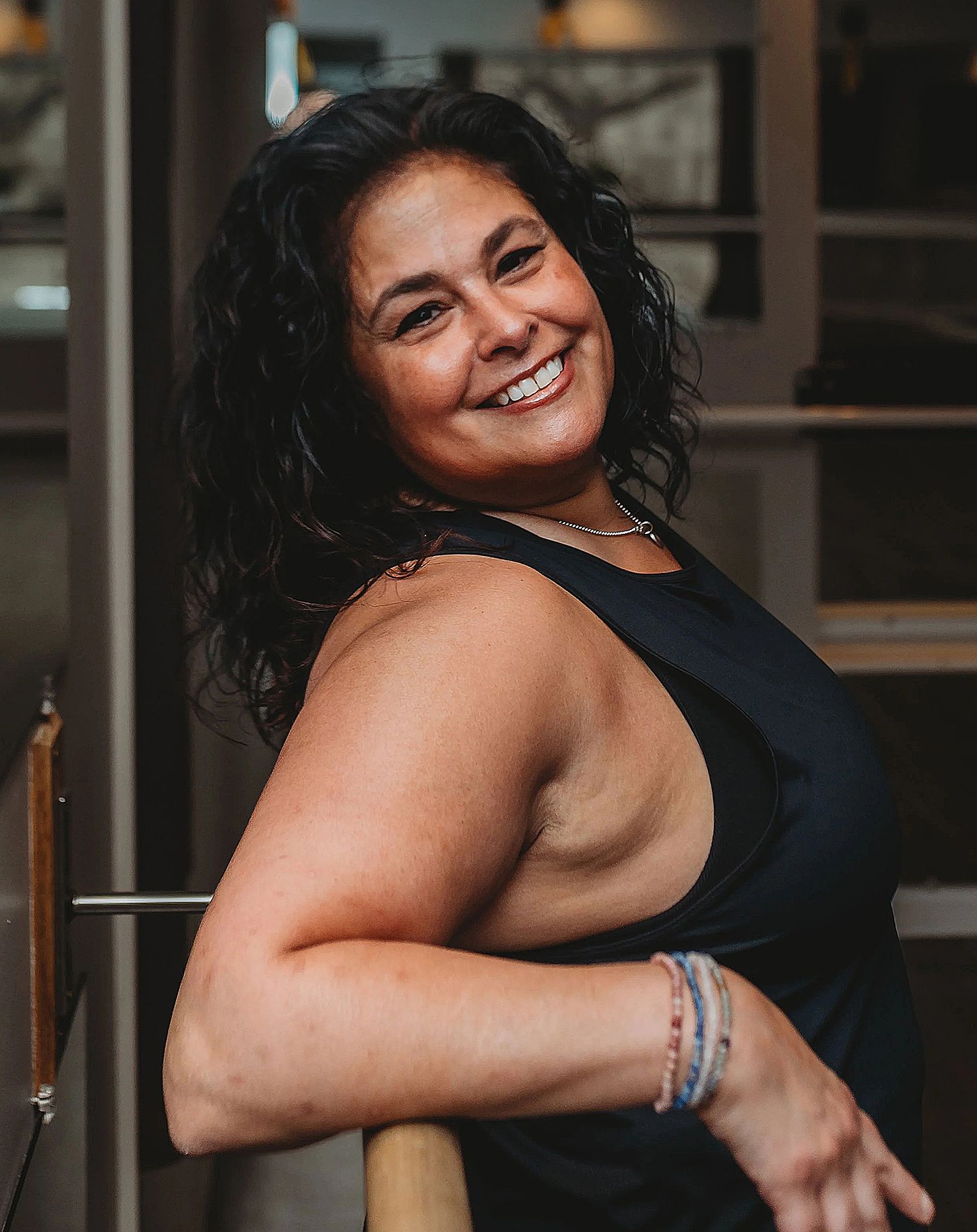 Woman smiling, leaning on a bar, wearing a black tank top, in a well-lit interior.