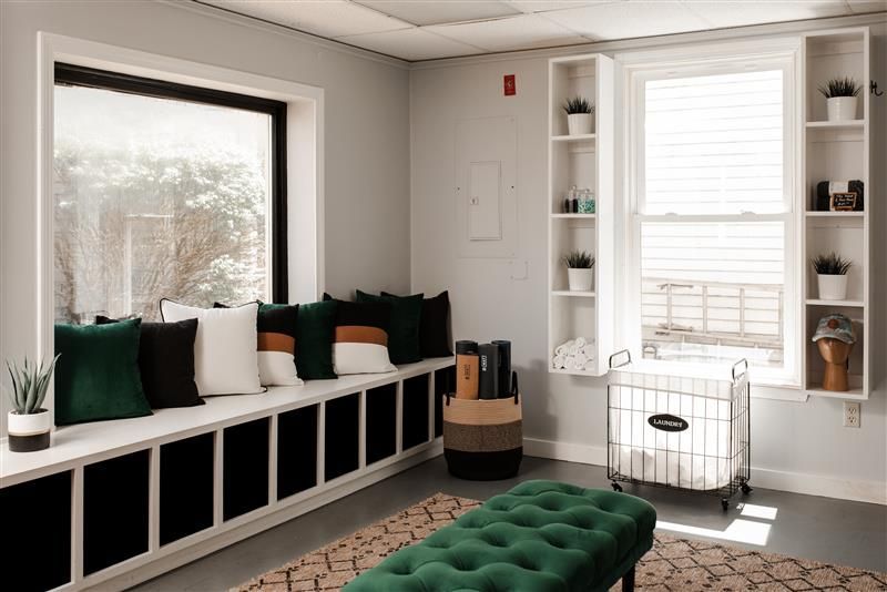 Cozy laundry room with window seat and shelving. Includes a green ottoman, laundry cart, and neutral rug.