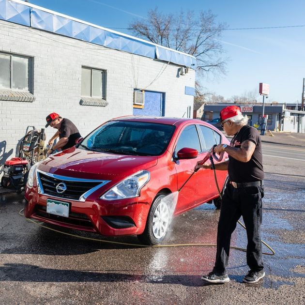 Two workers washing a red Nissan SUV outside a building on a sunny day.