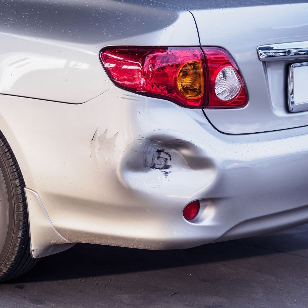 Scratched silver car rear bumper with a dent near the taillight.