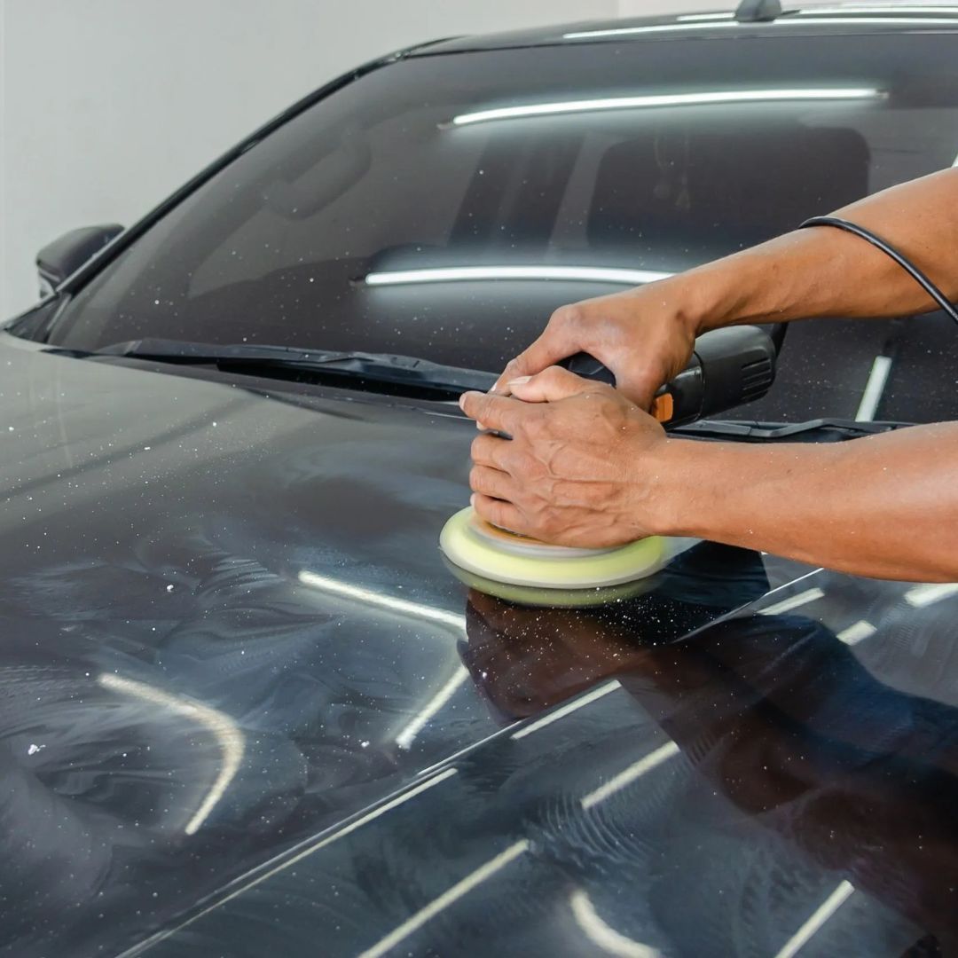 Hands buffing a black car hood with a polishing machine in a garage