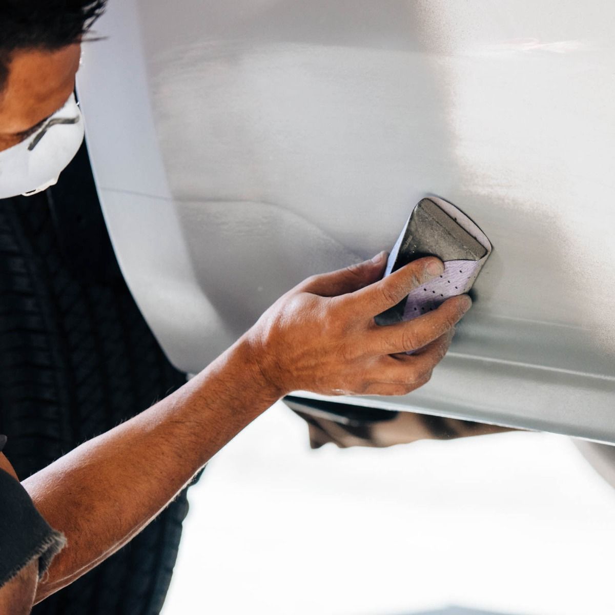 Person sanding a white car panel with a handheld tool in a workshop