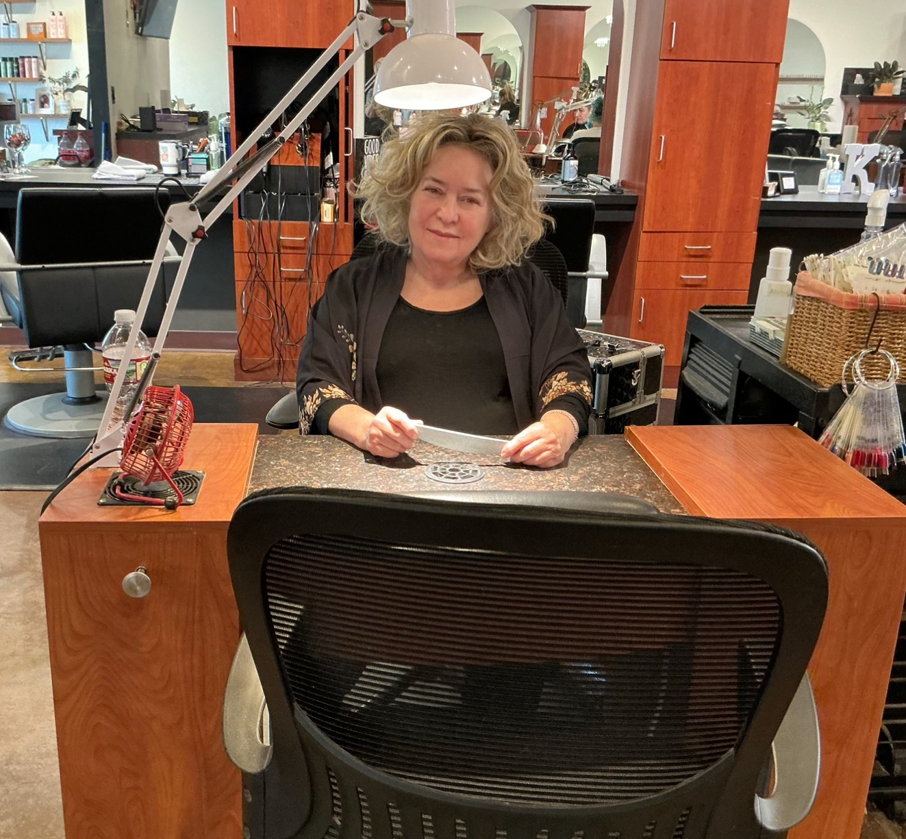 A woman with curly hair at a nail station in a salon, holding a paper.
