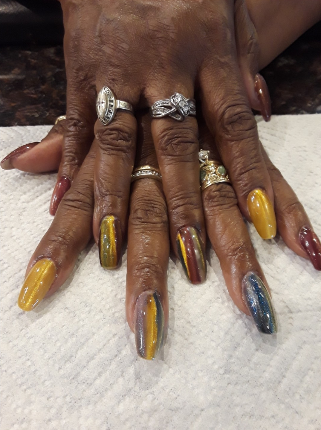 Hands with long, colorful nails. Rings adorn fingers. The nails are painted various fall colors and textures.