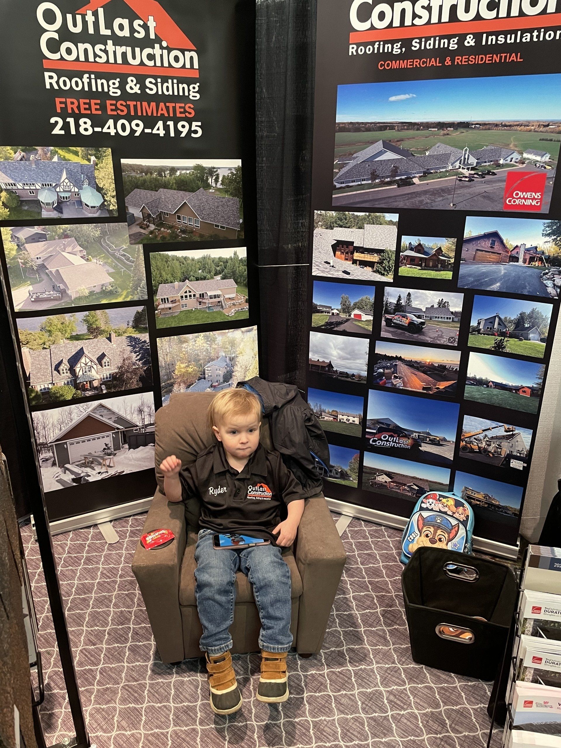 A little boy is sitting in a chair in front of a sign for outlast construction.
