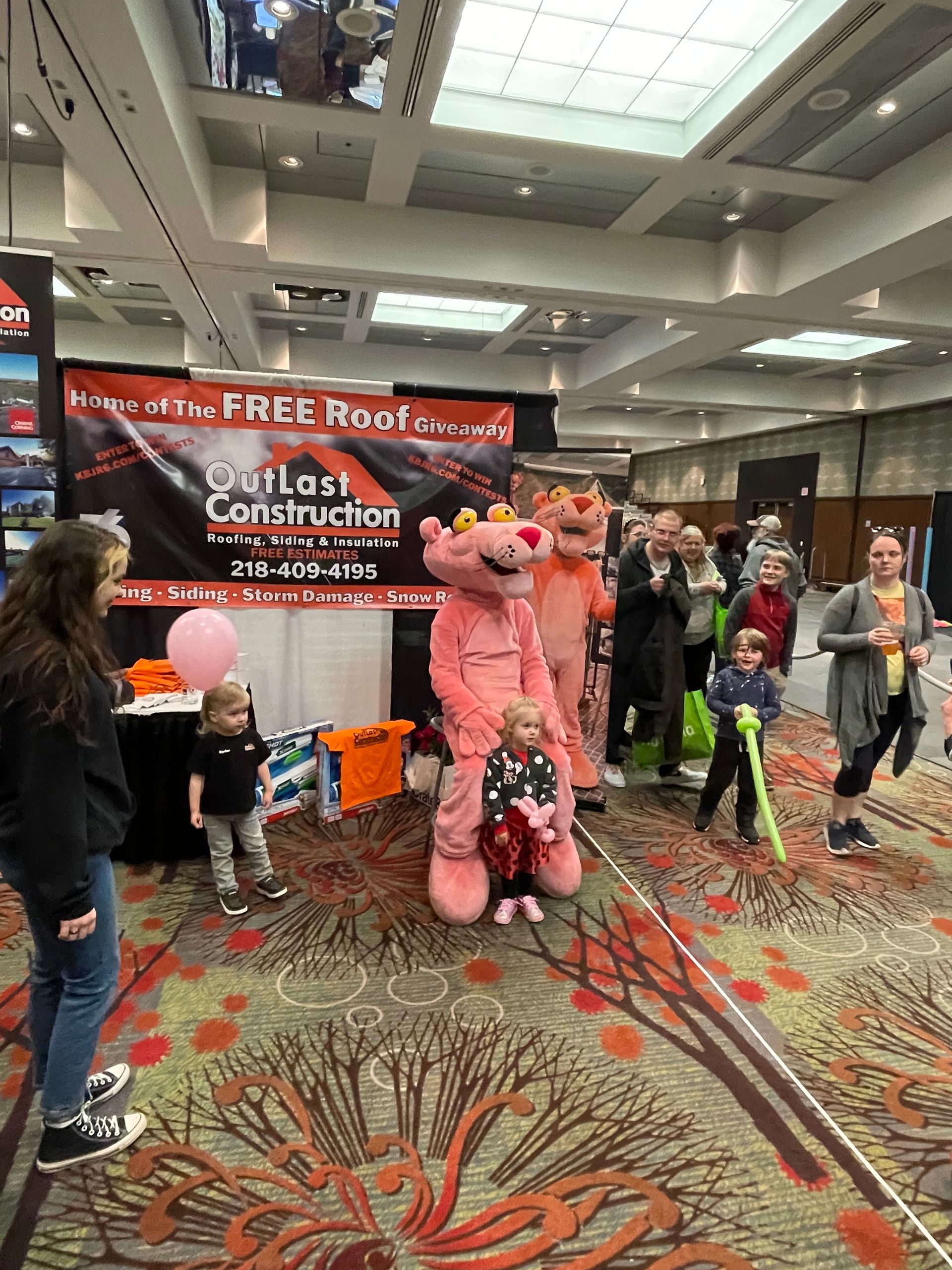 A group of people are standing around a pink panther mascot at a convention.
