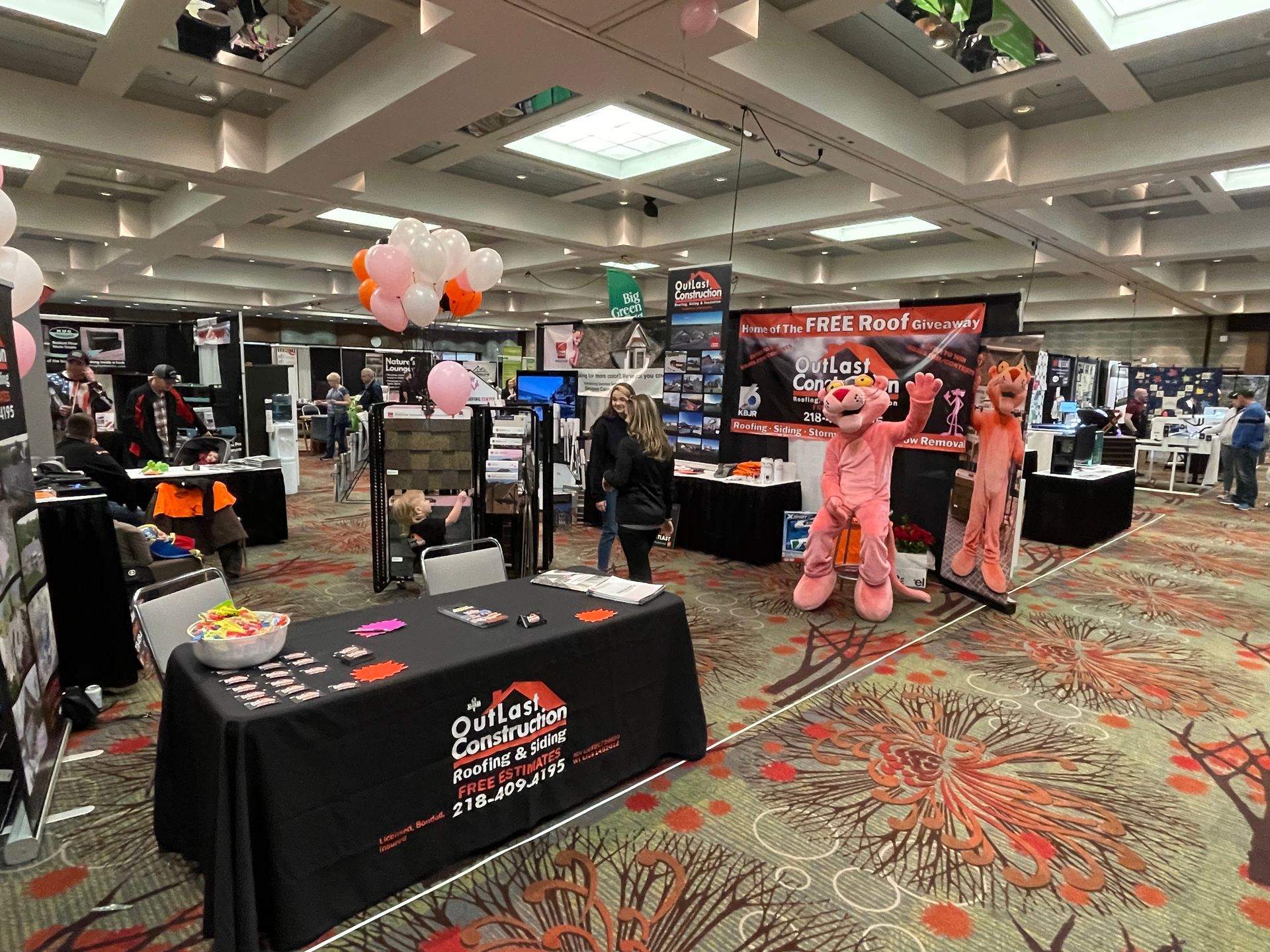 A large room filled with tables , balloons , and a pink mascot.
